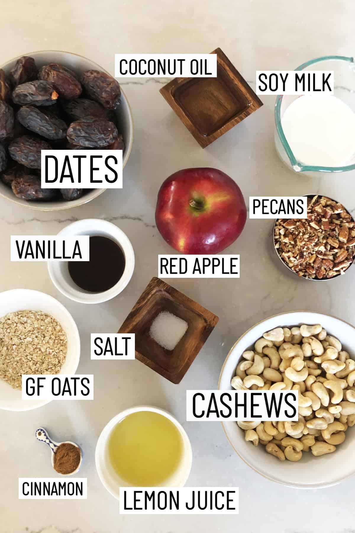 Flat lay image of portioned ingredients of raw vegan cheesecake including lemon juice, cinnamon, cashews, salt, oats, pecans, red apple, dates, vanilla, soy milk, and coconut oil.