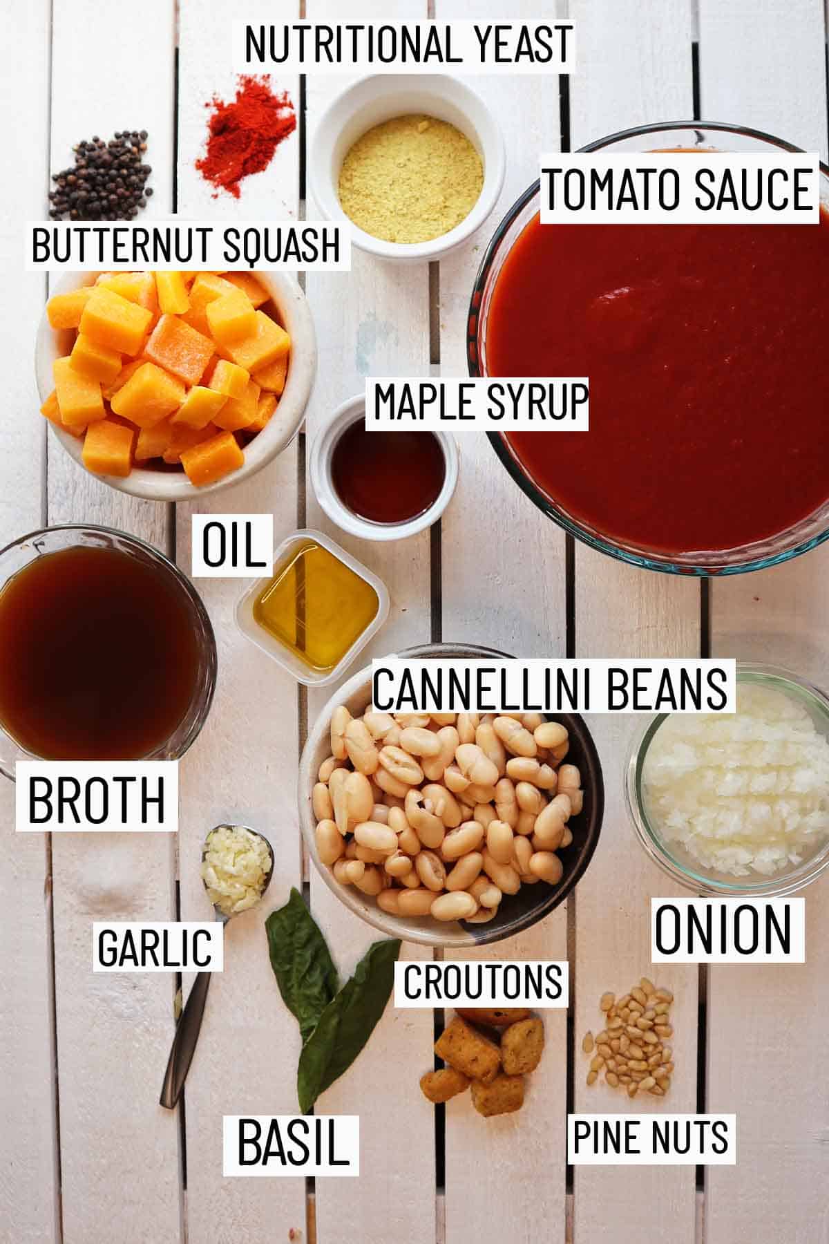 Flat lay image of portioned recipe ingredients including tomato sauce, broth, maple syrup, onion, garlic, white beans, butternut squash, nutritional yeast, basil, and croutons. 