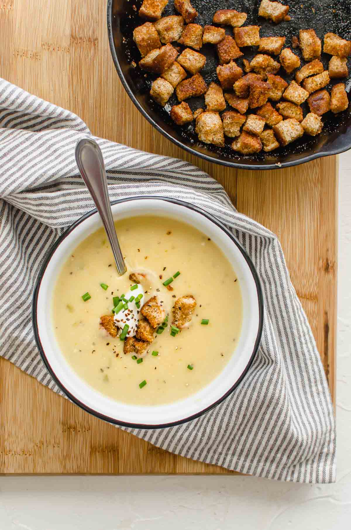 Birds eye view of vegan potato soup in a bowl topped with croutons and chives, with more croutons on a pan in the background. 