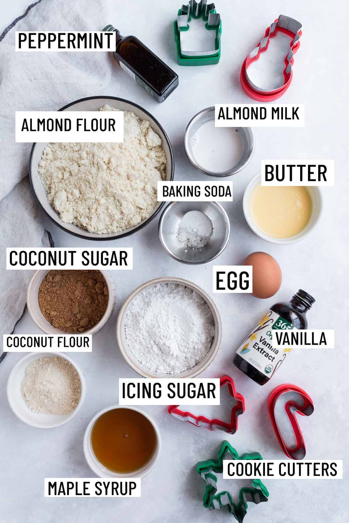 Birds eye view image of portioned ingredients for gluten free Christmas cookie recipe including almond flour, vanilla and peppermint extract, butter, baking soda, almond milk, egg, coconut sugar, icing sugar, coconut flour, maple syrup, and cookie cutters. 