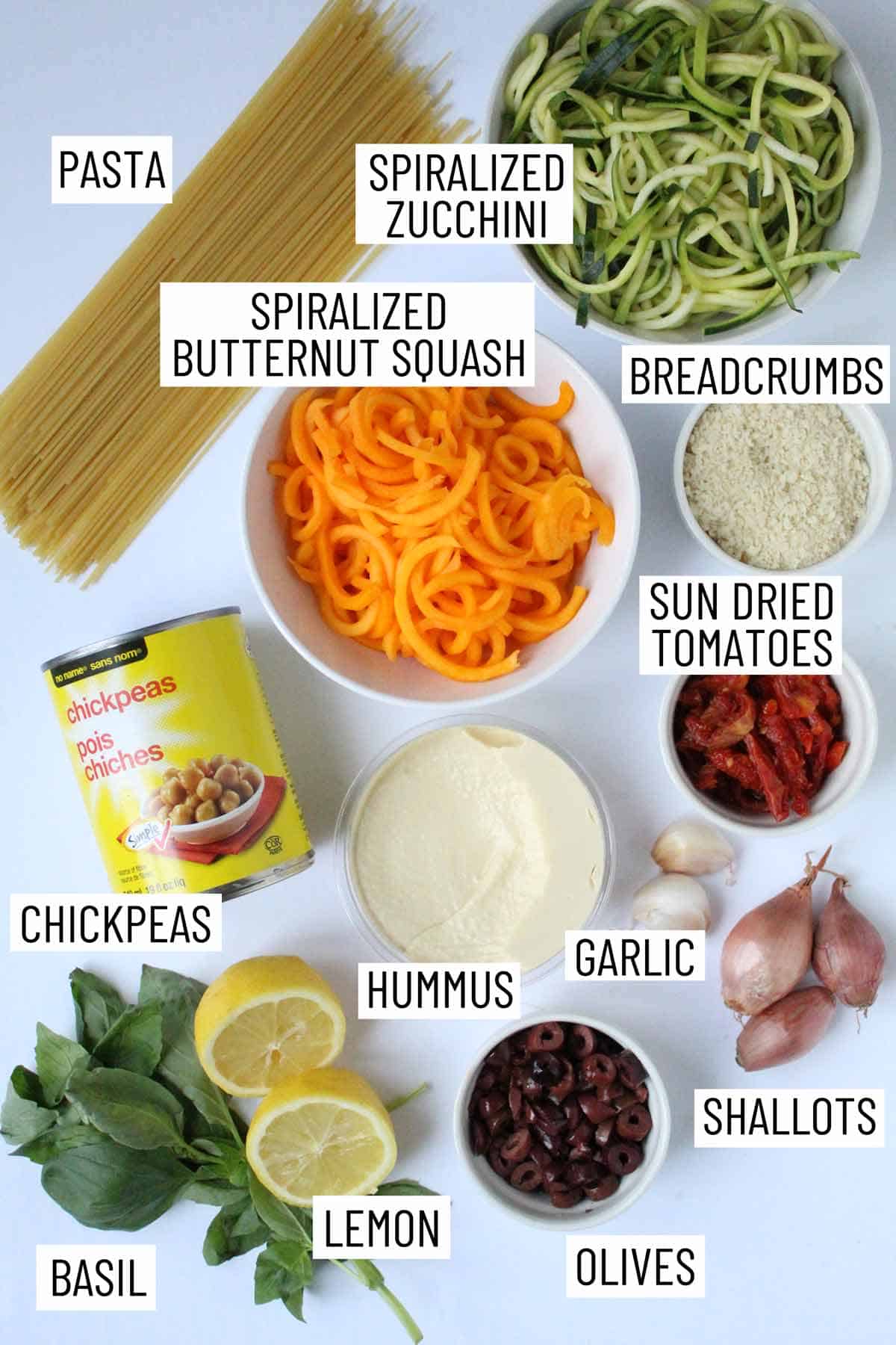 Overhead photo of ingredients for hummus pasta including basil, lemon, olives, shallots, garlic, hummus, chickpeas, sun dried tomatoes, breadcrumbs, spiralized butternut squash, spiralized zucchini, and pasta.