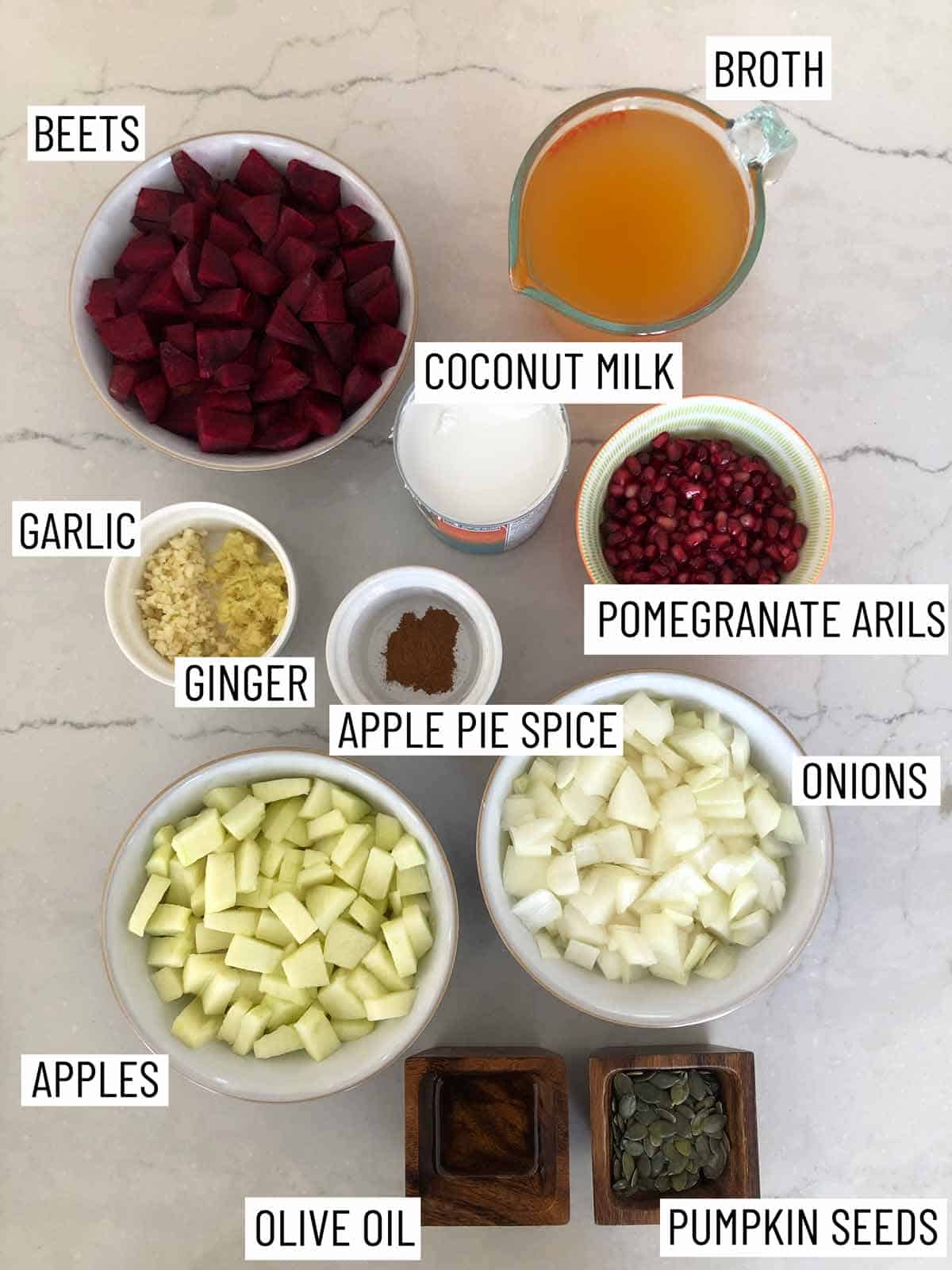 Overhead image of ingredients: broth, coconut milk, pomegranate arils, onions, pumpkin seeds, olive oil, apples, apple pie spice, ginger, garlic, and beets.