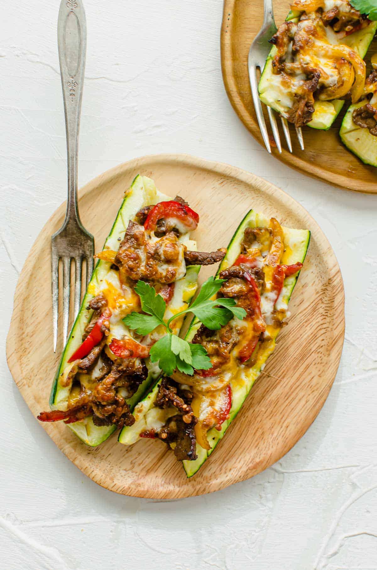 Birds eye view of prepared zucchini boats with Philly cheesesteak stuffing on a wooden plate topped with parsley.