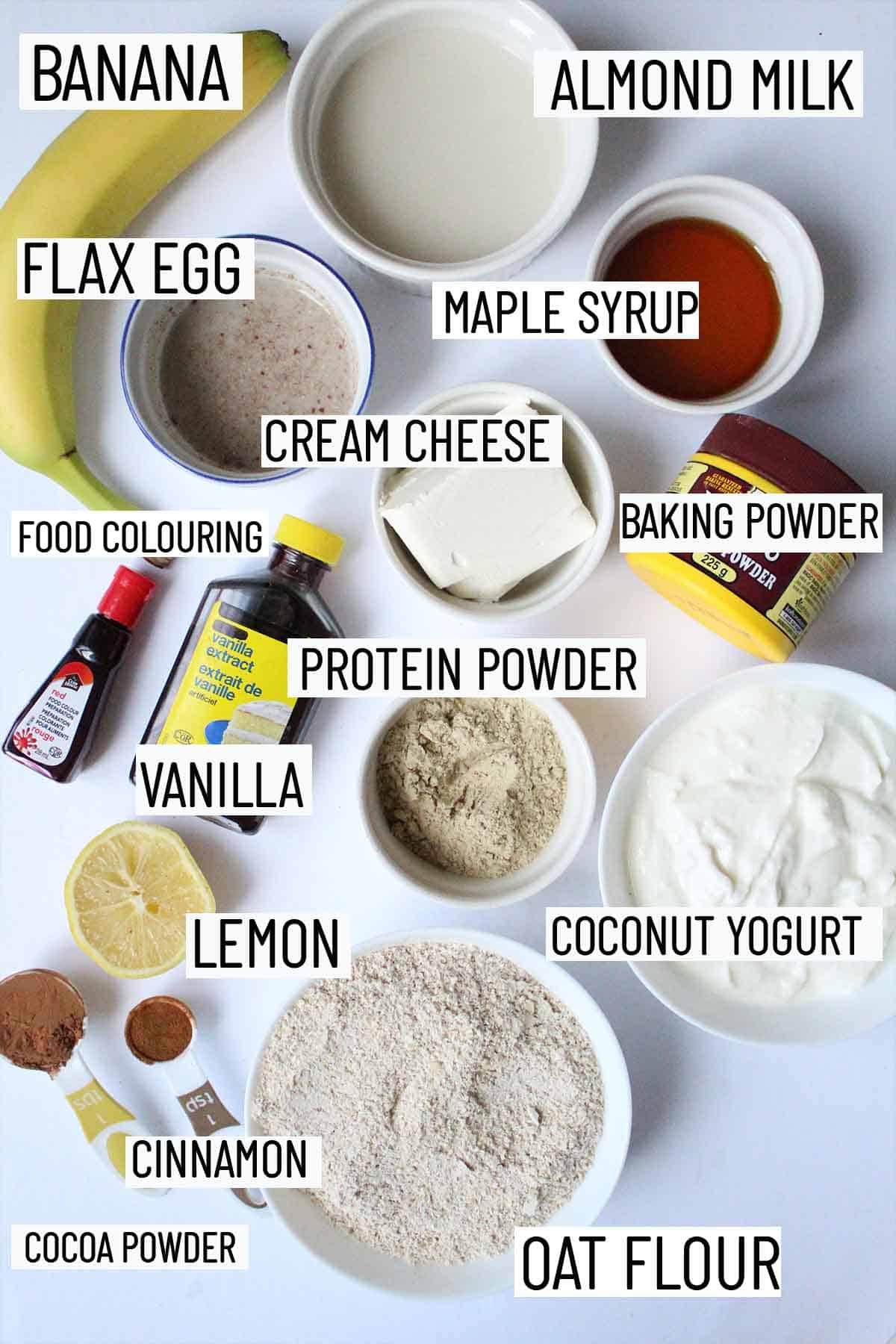 Flat lay image of recipe ingredients including oat flour, cocoa powder, cinnamon, lemon, protein powder, vanilla, food colouring, cream cheese, baking powder, maple syrup, banana, flax egg and almond milk. 