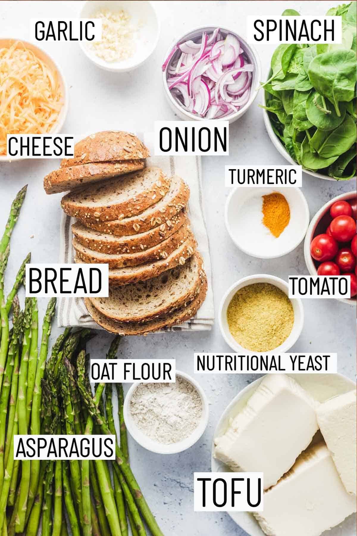 Flat lay image of recipe ingredients including bread, spinach, onion, garlic, cheese, oat flour, tofu, asparagus, nutritional yeast, tomato, and turmeric.