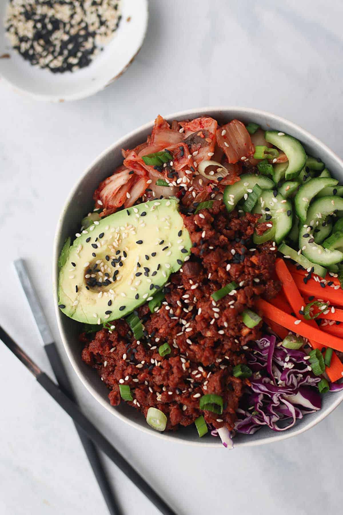 Birds eye view of Korean bowl with veggie toppings and vegan meat garnished with sesame seed and green onion with two chop sticks laying on the side. 