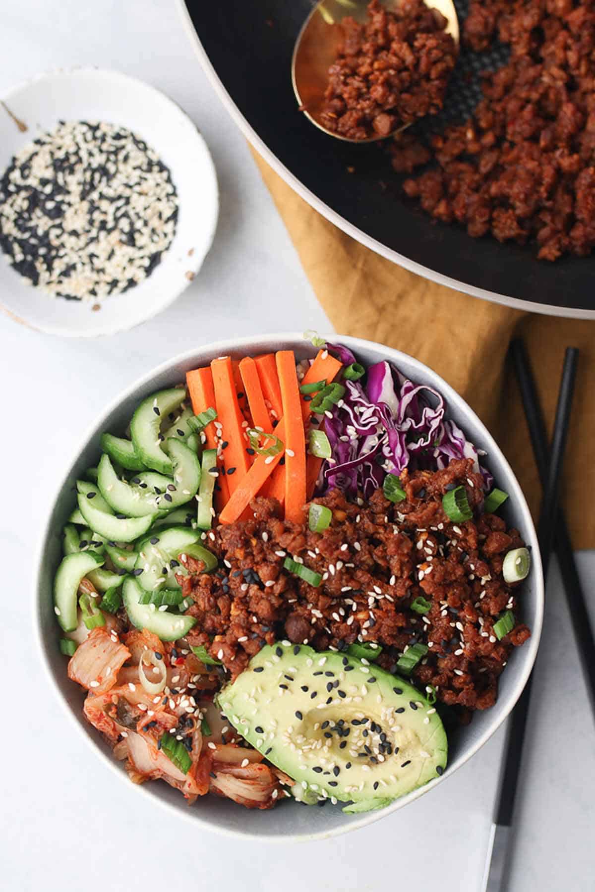 Birds eye view of vegan buddha bowl garnished with green onion and sesame seed with a bowl of sesame seeds in the background and two chopsticks laying on the side. 