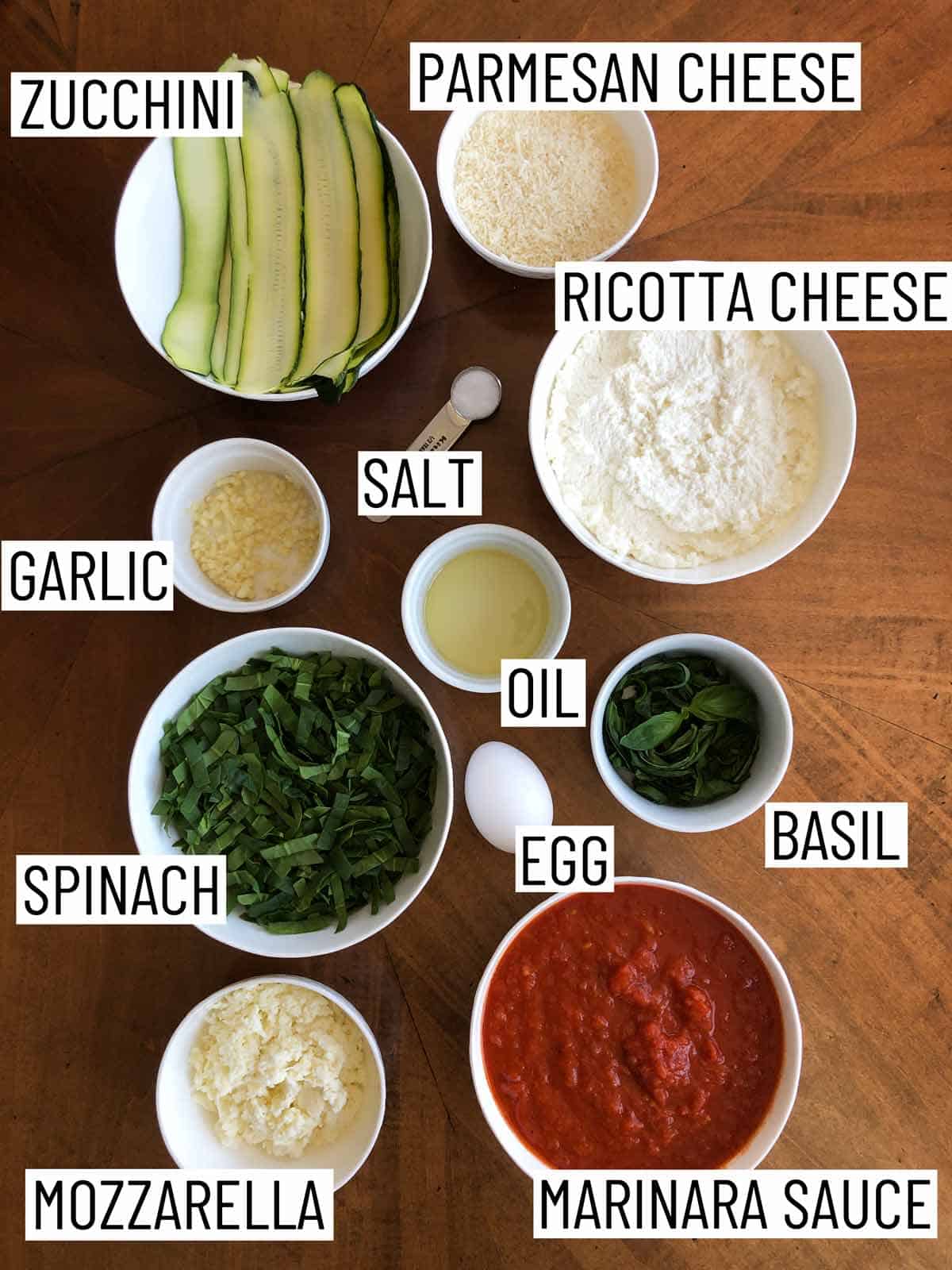 Overhead image of ingredients: zucchini, parmesan cheese, ricotta cheese, salt, oil, basil, egg, marinara sauce, mozzarella, spinach, and garlic.