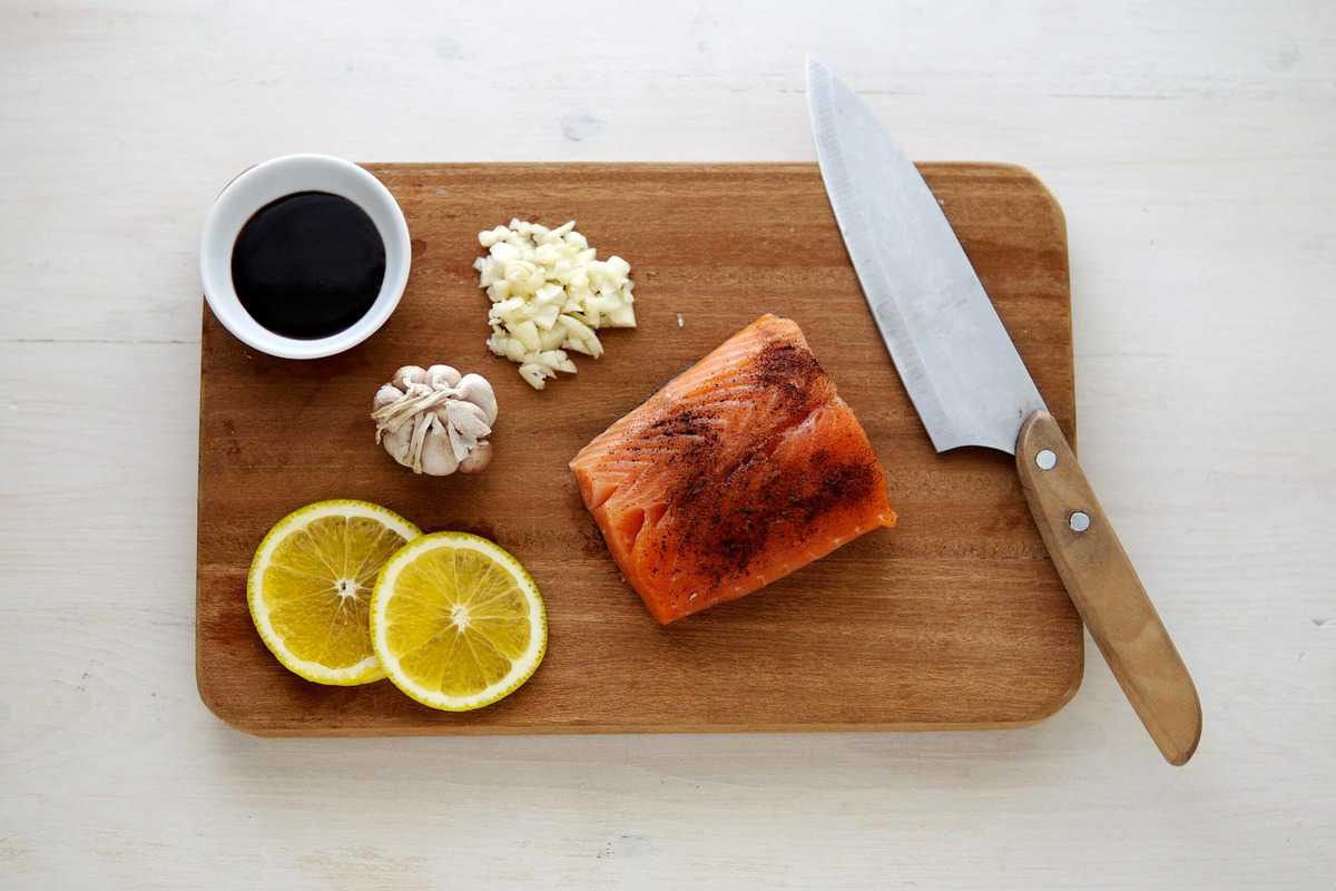 A piece of seasoned salmon, lemon slices, garlic, a chefs knife and soy sauce on a cutting board. 