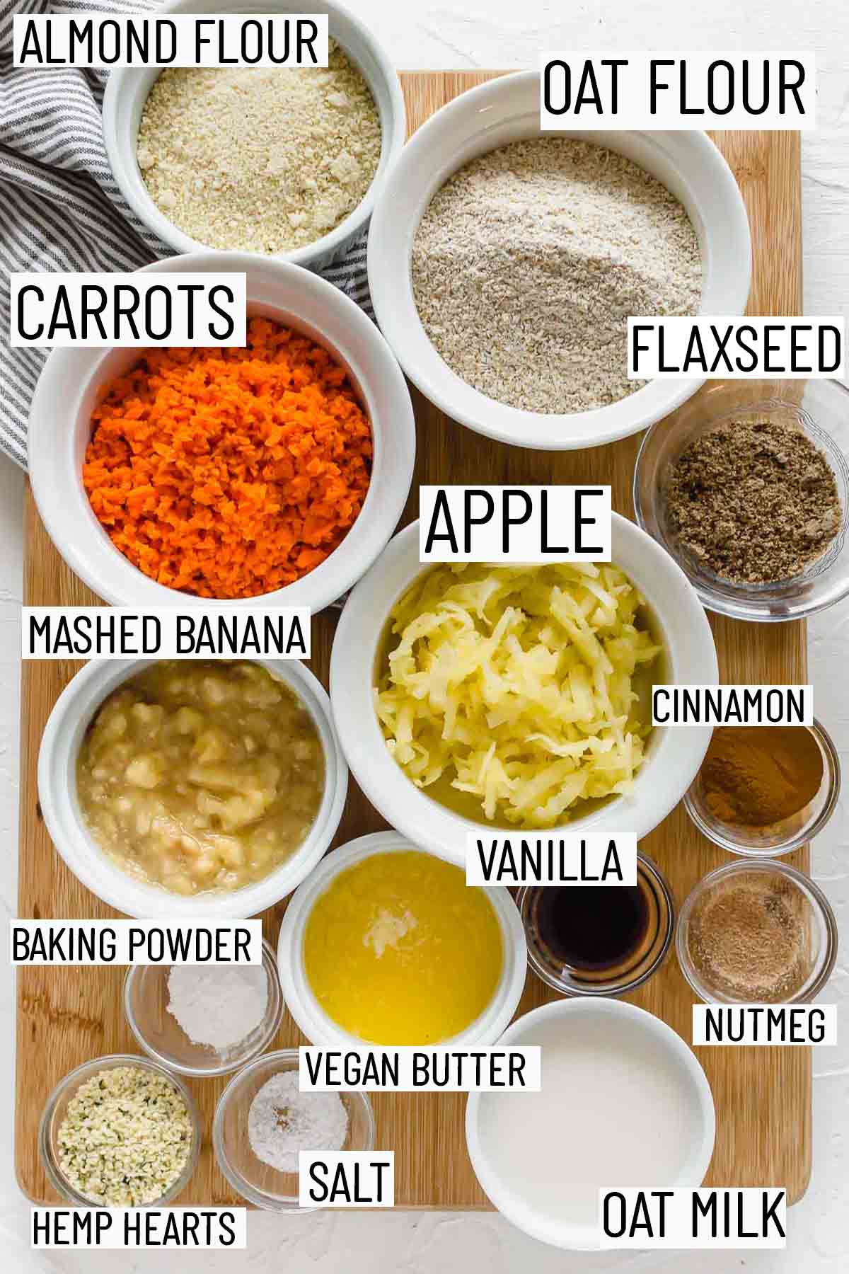 Flat lay image showing portioned recipe ingredients including oat flour, almond flour, apple, carrot, banana, baking powder, salt, hemp hearts, oat milk, nutmeg, vanilla, cinnamon, and flax seed.