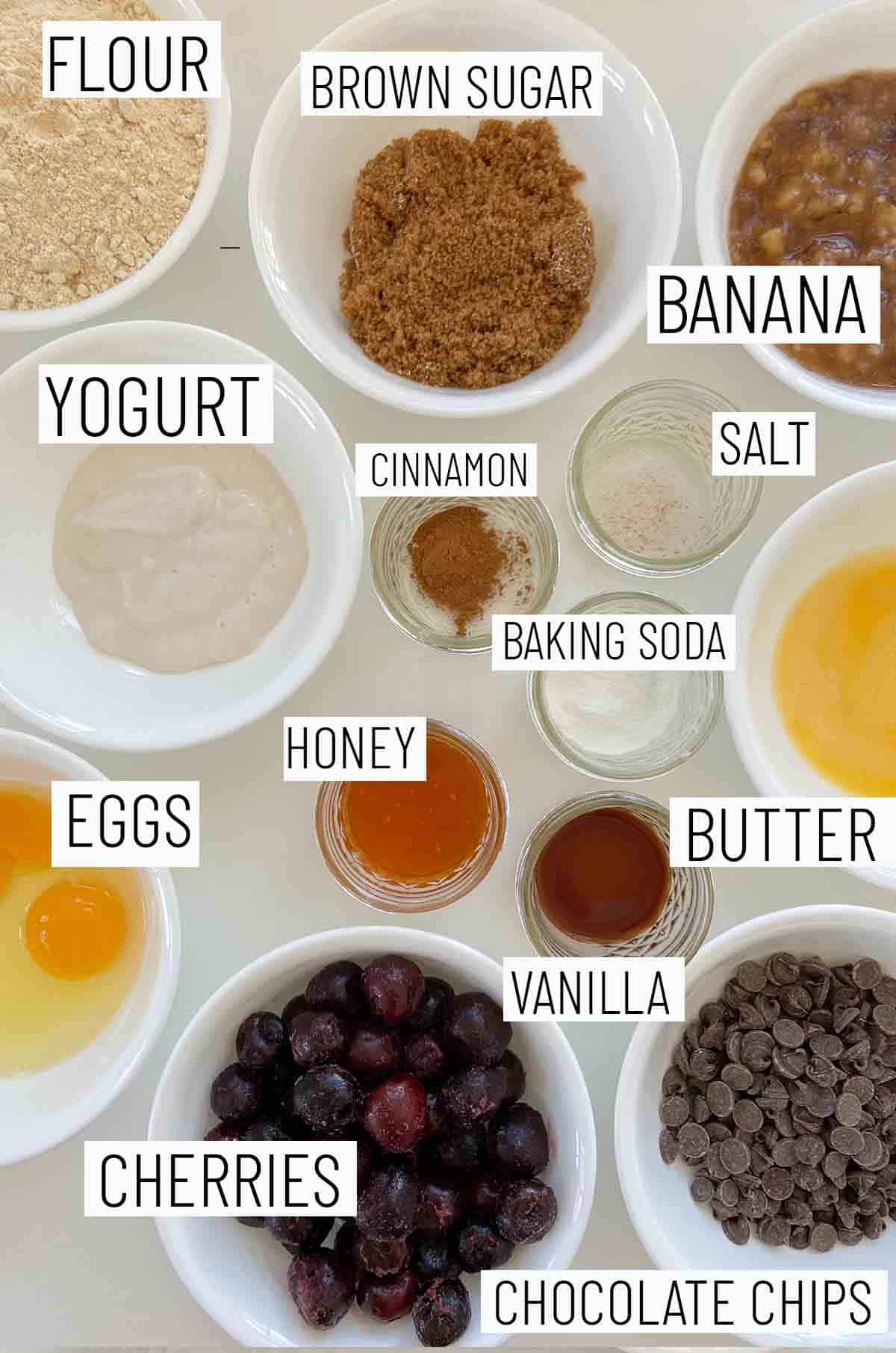 Flat lay image of portioned recipe ingredients including chocolate chips, cherries, eggs, butter, vanilla, honey, baking soda, salt, cinnamon, yogurt, banana and flour.