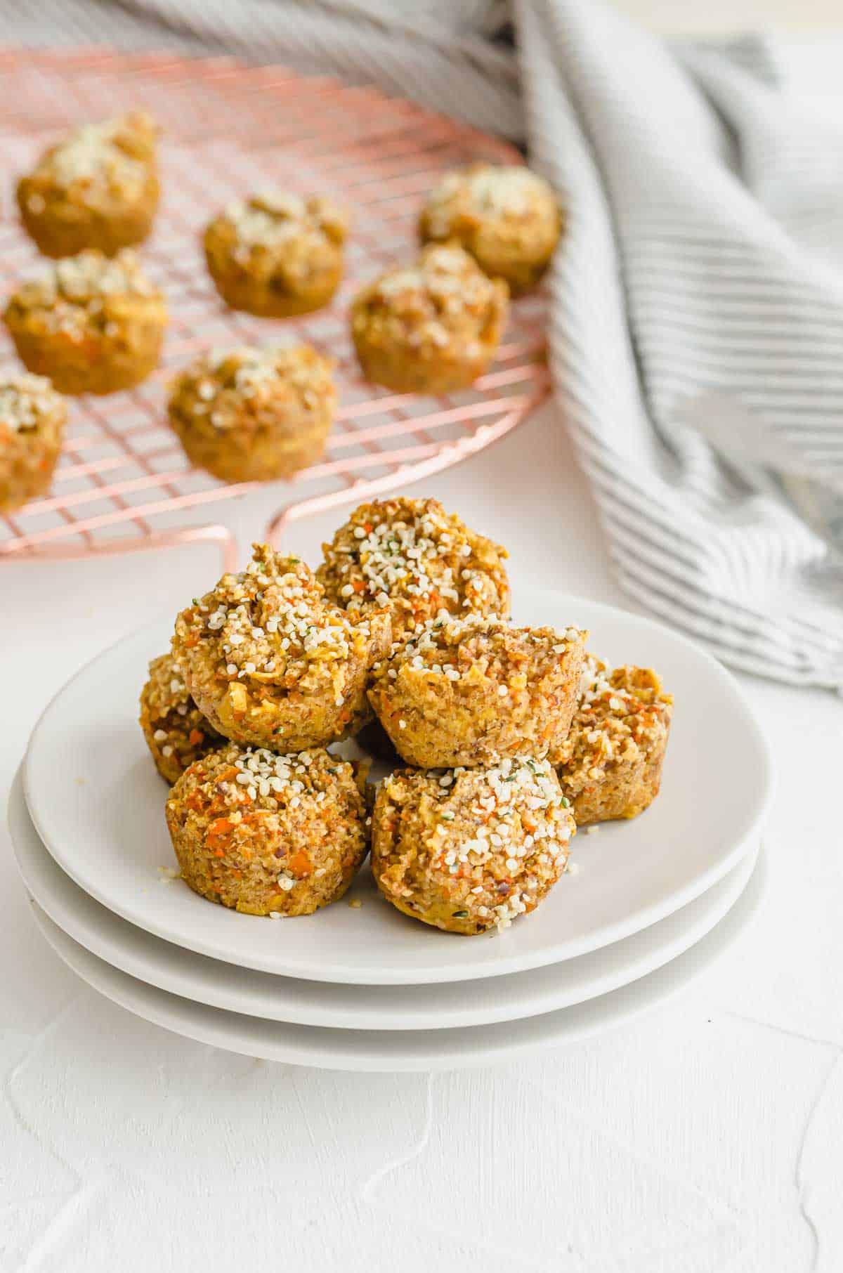 Several baby carrot muffins on a white plate with more cooling in a cooling rack in the background. 