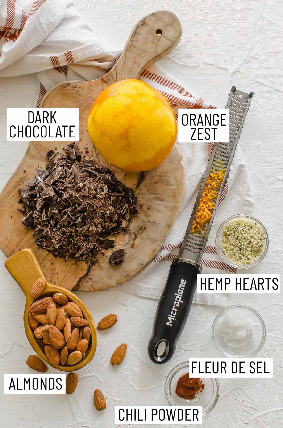 Overhead photo of ingredients needed to make chocolate covered almonds recipe: dark chocolate, orange zest, hemp hearts, fleur de sel, chili powder, and almonds.