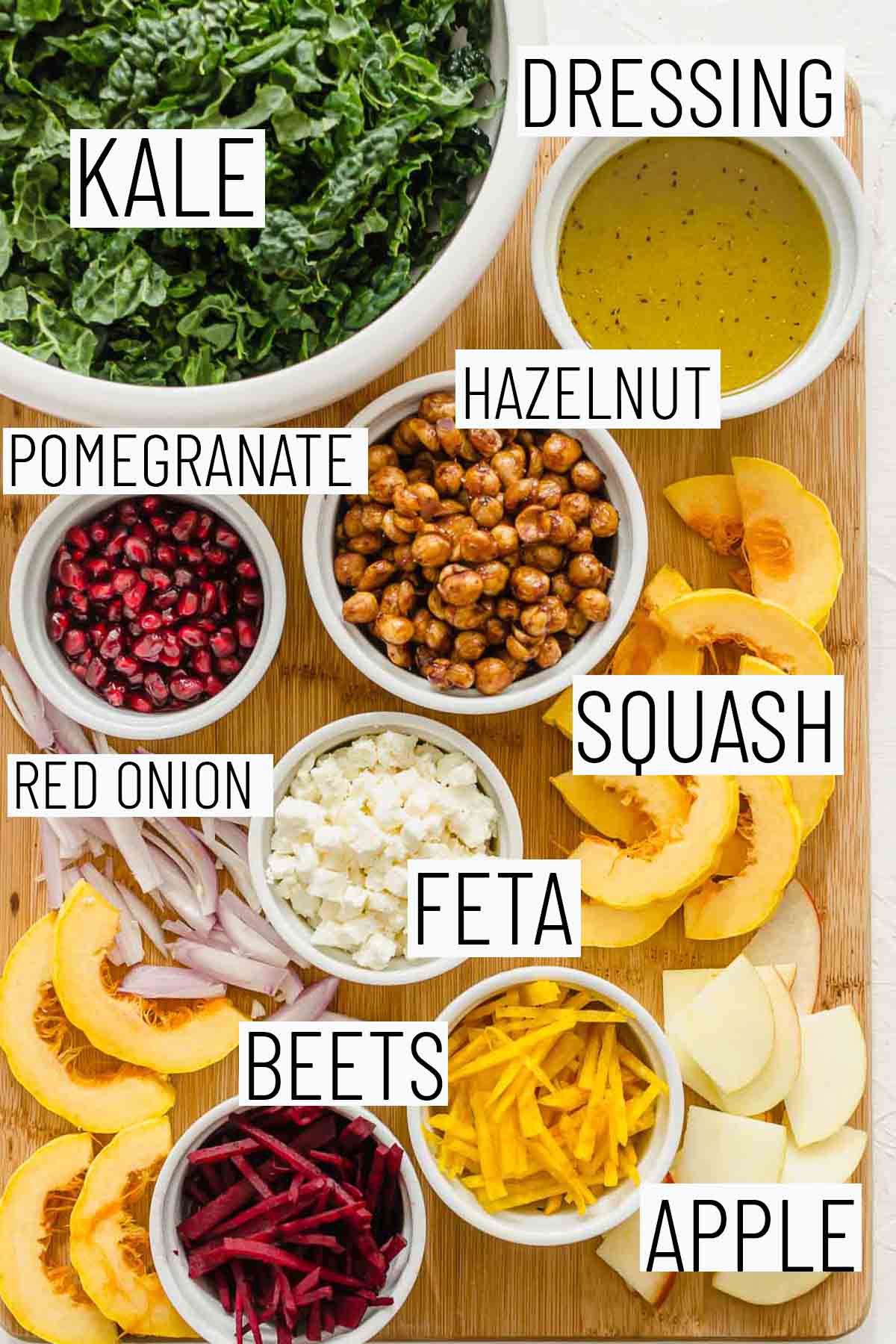 Flat lay image of recipe ingredients including kale, squash, beets, apple, red onion, pomegranate arils, hazelnut, and salad dressing. 