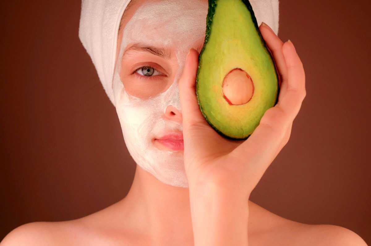 Woman with a white face mask holding up an avocado to her eye.  