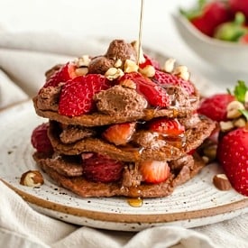 A stack of nutella pancakes with strawberries and syrup being drizzled on.