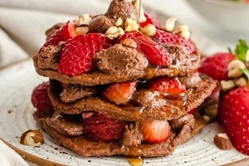 A stack of nutella pancakes with strawberries and syrup being drizzled on.