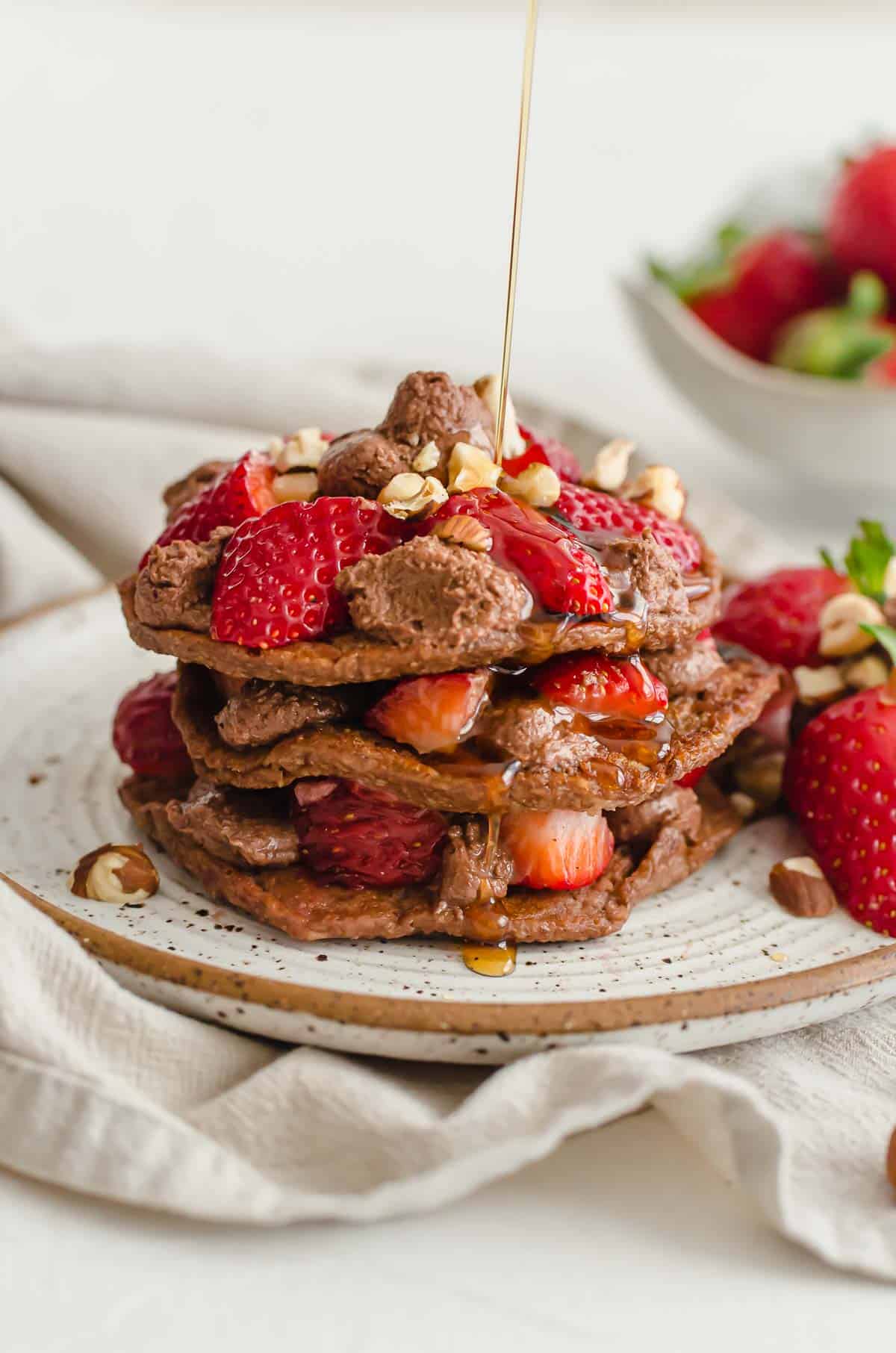 A stack of nutella pancakes with strawberries and syrup being drizzled on.