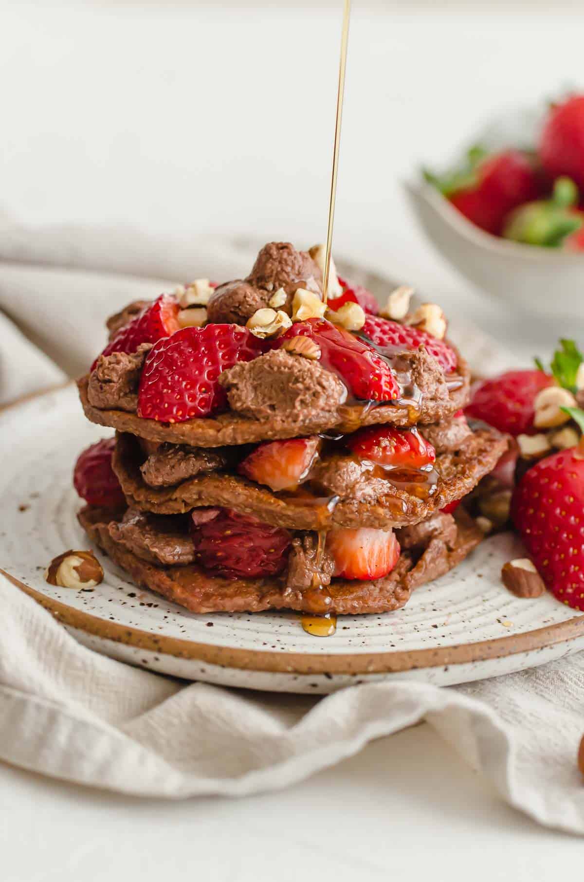 A stack of nutella stuffed pancakes with strawberries and syrup being drizzled on.