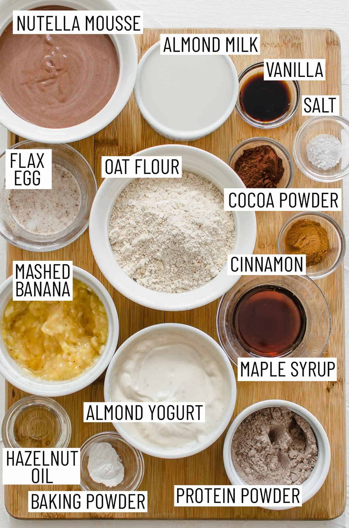 Overhead image of ingredients needed to make pancakes: mousse, almond milk, vanilla, salt, cocoa powder, cinnamon, oat flour, flax egg, mashed banana, maple syrup, almond yogurt, hazelnut oil, baking powder, and protein powder.