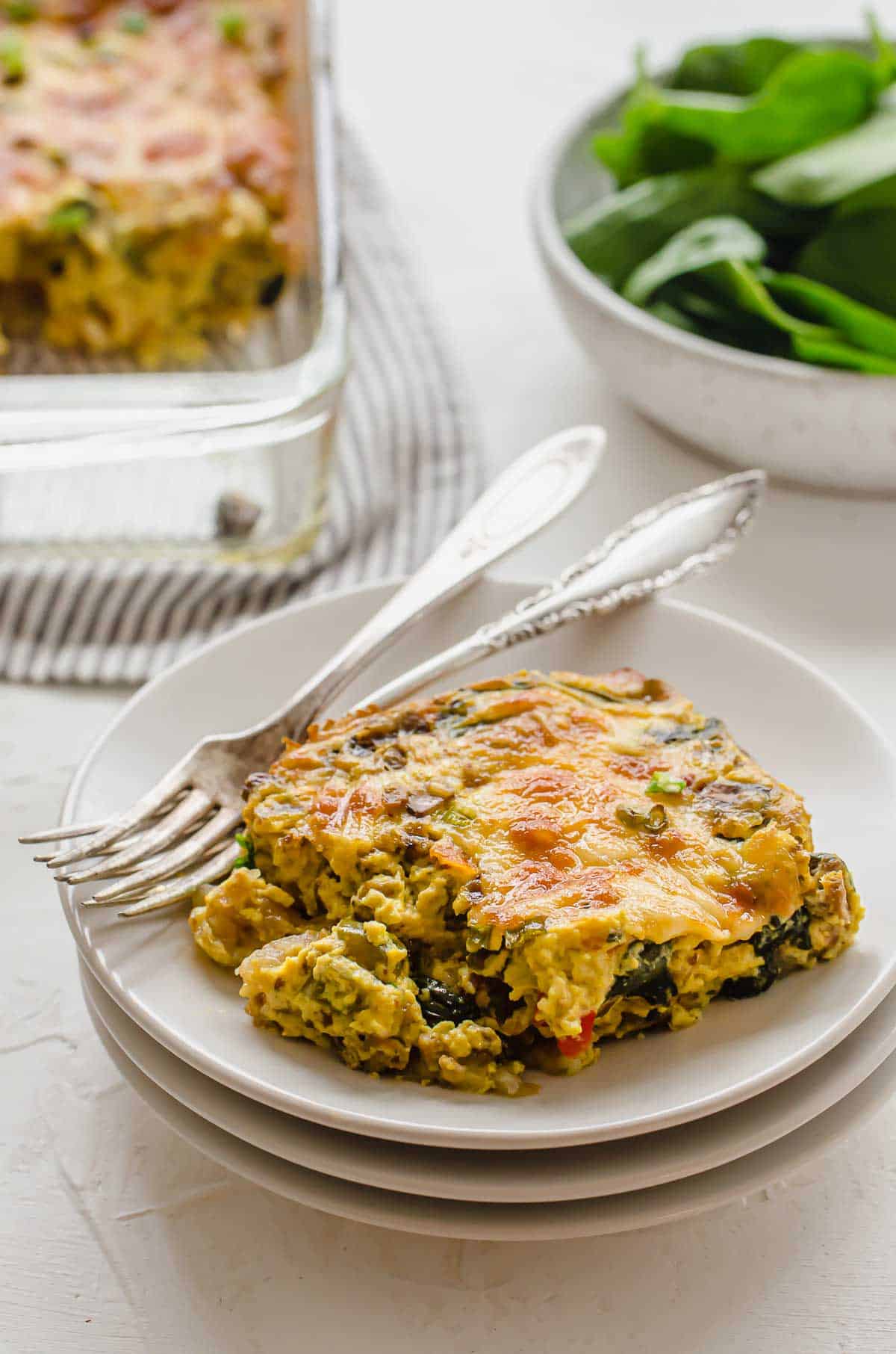 Close up of breakfast casserole on three stacked white plates with two forks on the side with a bowl of spinach and the remaining casserole in the background. 