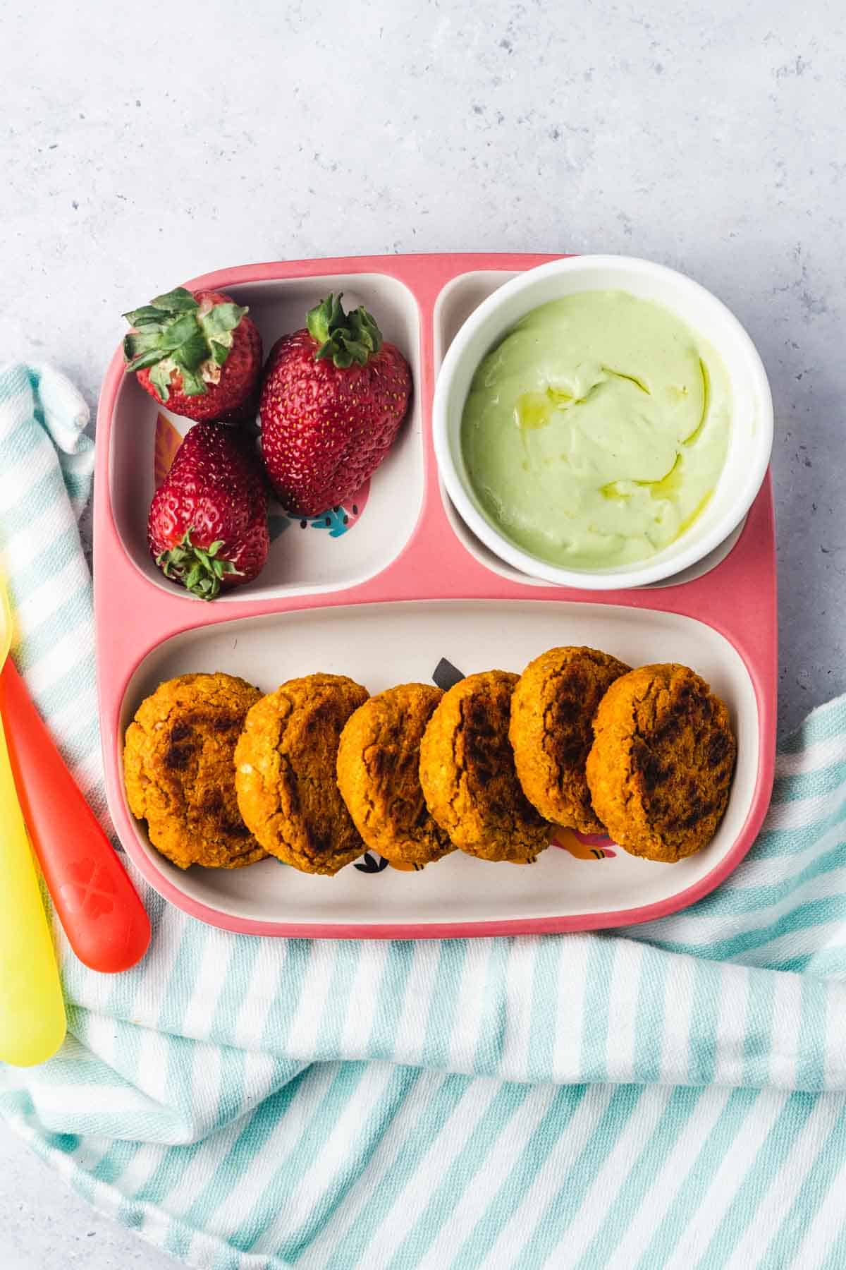 Birds eye view of plated sweet potato chickpea patties in a feeding tray served with avocado sauce and strawberries. 