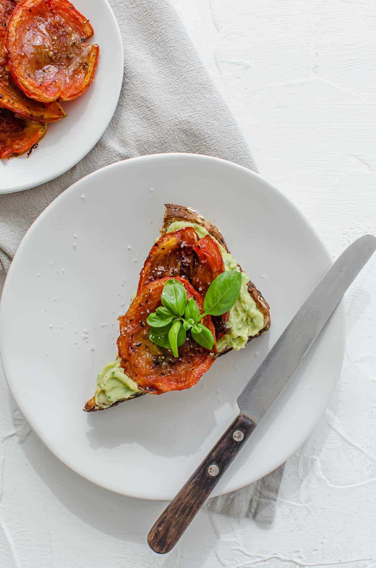 Birds eye view of vegan avocado toast on a white plate topped with roasted tomatoes and garnished with fresh basil. 