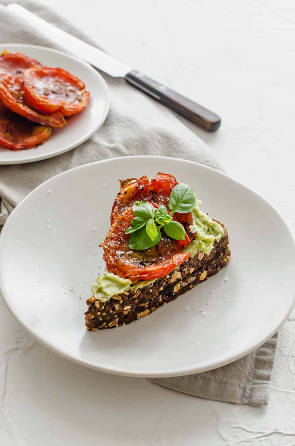 Close up of vegan avocado toast on a white plate topped with roasted tomato and a basil leaf with extra roasted tomatoes in the background.