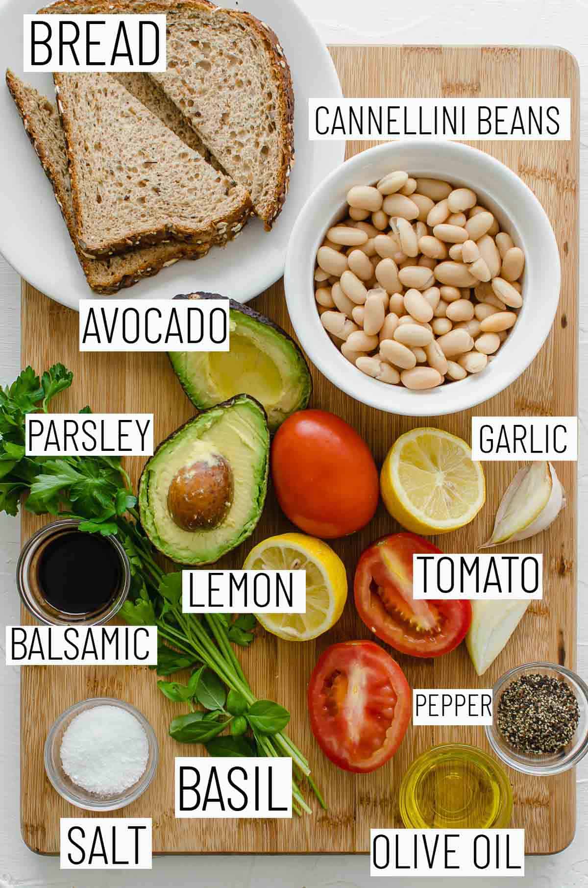 Flat lay image of recipe ingredients including lemon, pepper, Roma tomatos, olive oil, salt, basil, balsamic, parsley, white bean, avocado, and bread. 