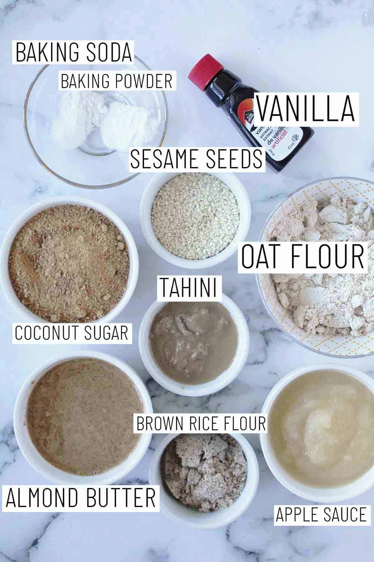 Flat lay image showing portioned recipe ingredients including oat flour, tahini, coconut sugar, brown rice flour, almond butter, apple sauce, vanilla, sesame seeds, baking soda, and baking powder. 