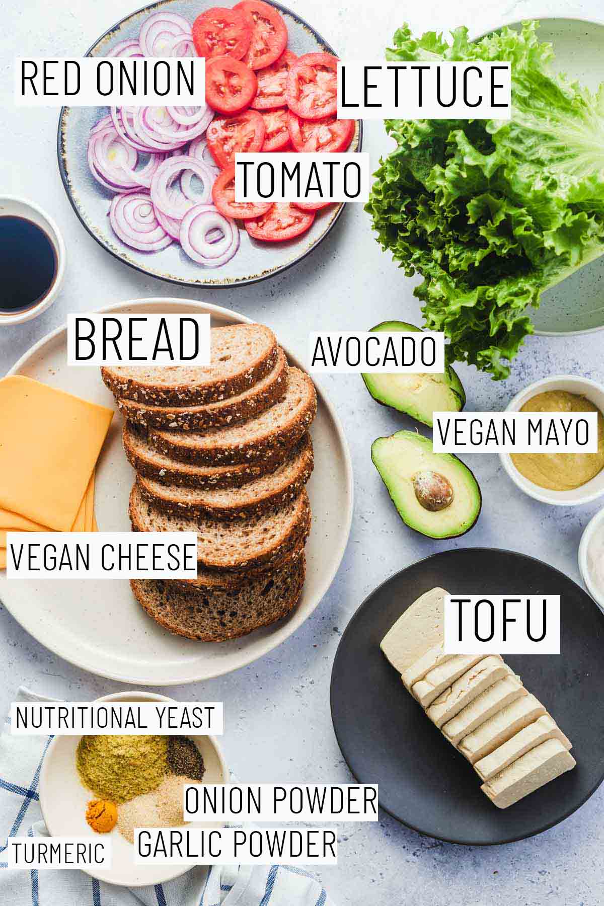 Flat lay image of portioned recipe ingredients including bread, cheese, mayo, tofu, lettuce, tomato, red onion, and seasonings. 