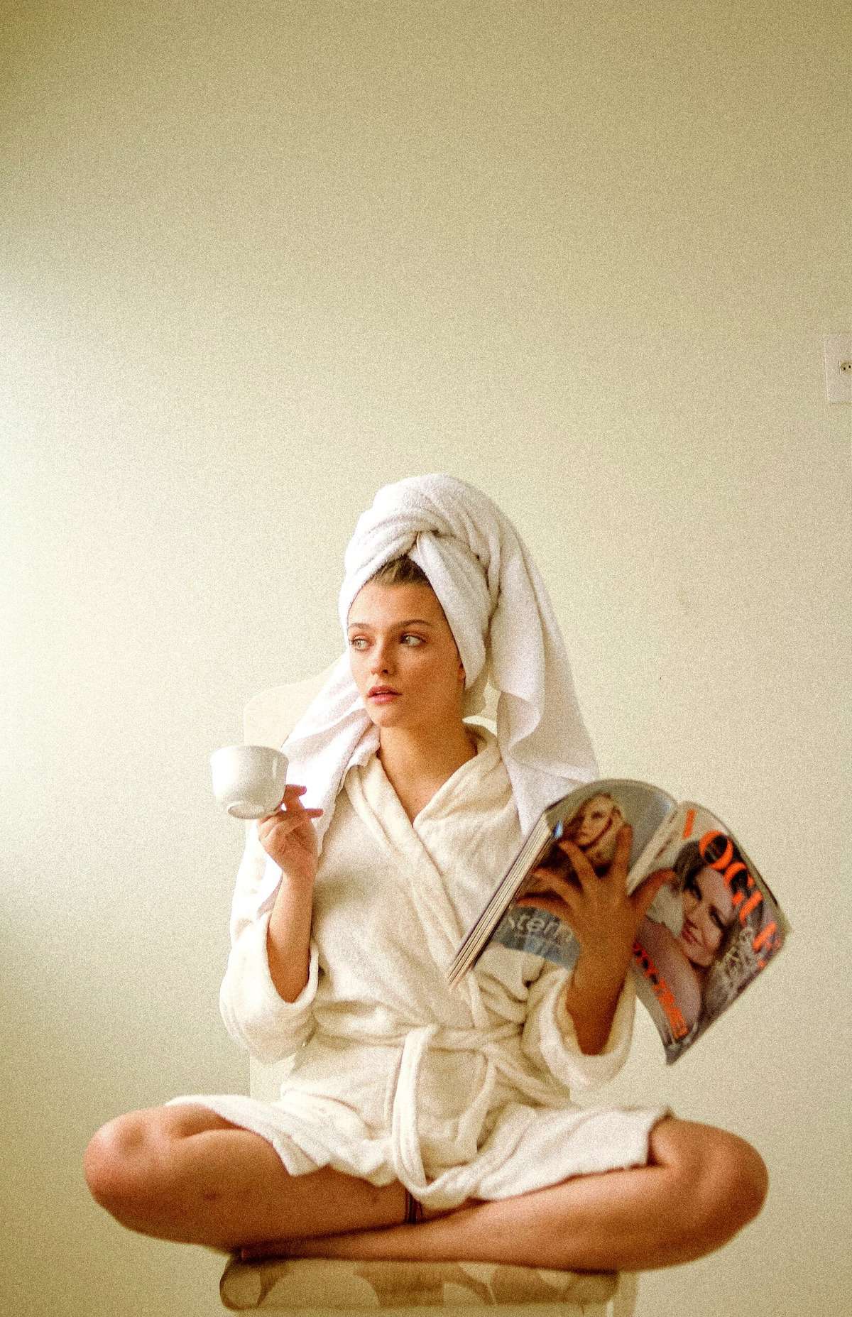 A woman in a robe, drinking coffee, and reading a magazine. 