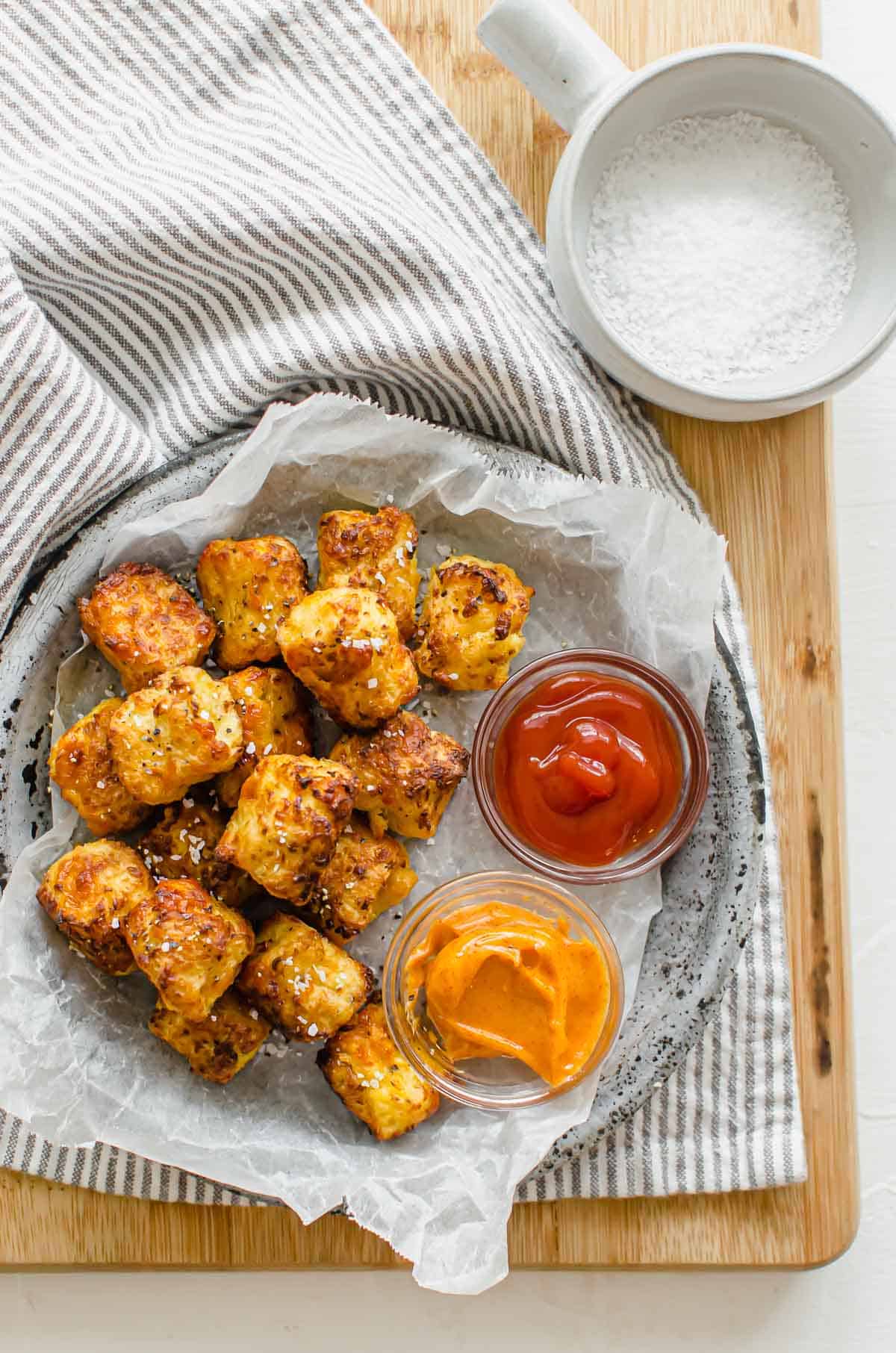 Birds eye view of cauliflower tater tots in a bowl with a side of ketchup and honey mustard sauce.