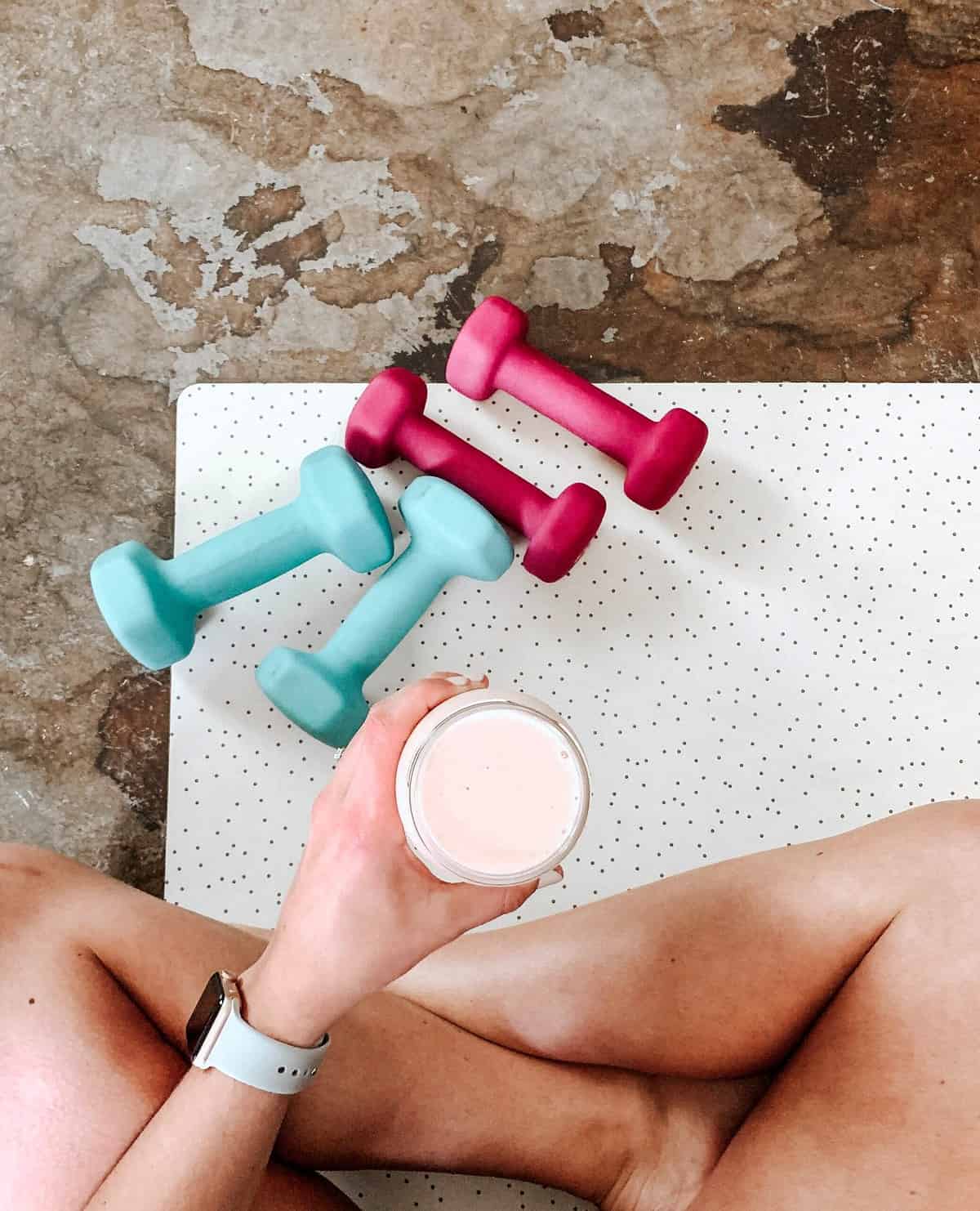 A person sitting on a yoga matt with free weights and holding up a protein smoothie. 