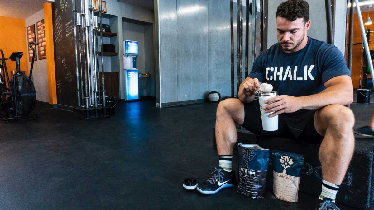 A man at the gym sitting on a bench and scooping protein powder into a tumbler mug. 