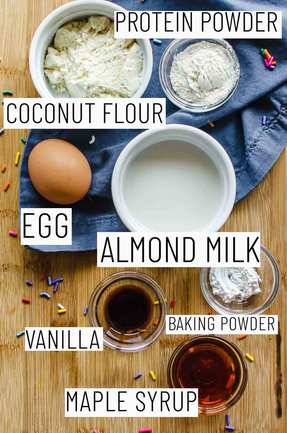 Flat lay image of portioned recipe ingredients including baking powder, maple syrup, vanilla, egg, coconut flour, and protein powder. 