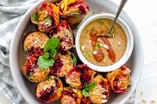 Overhead view of a white bowl of vegetable spring rolls cut in half nestled beside a bowl of homemade cashew sauce with a spoon inside.