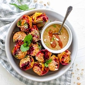 Overhead view of a white bowl of vegetable spring rolls cut in half nestled beside a bowl of homemade cashew sauce with a spoon inside.