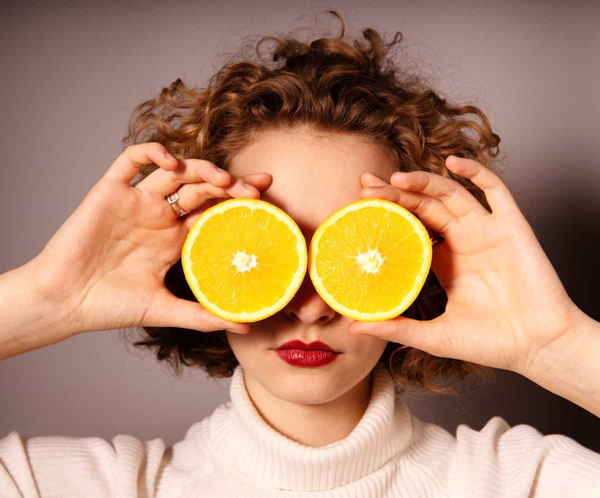 Woman holding up two orange slices up to her eyes. 