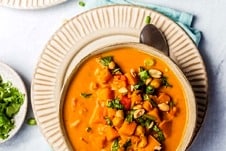 Overhead view of a bowl of sweet potato peanut soup on a plate with a spoon tucked under. Pinch bowl of garnishes off to the side.