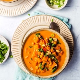 Overhead view of a bowl of sweet potato peanut soup on a plate with a spoon tucked under. Pinch bowl of garnishes off to the side.