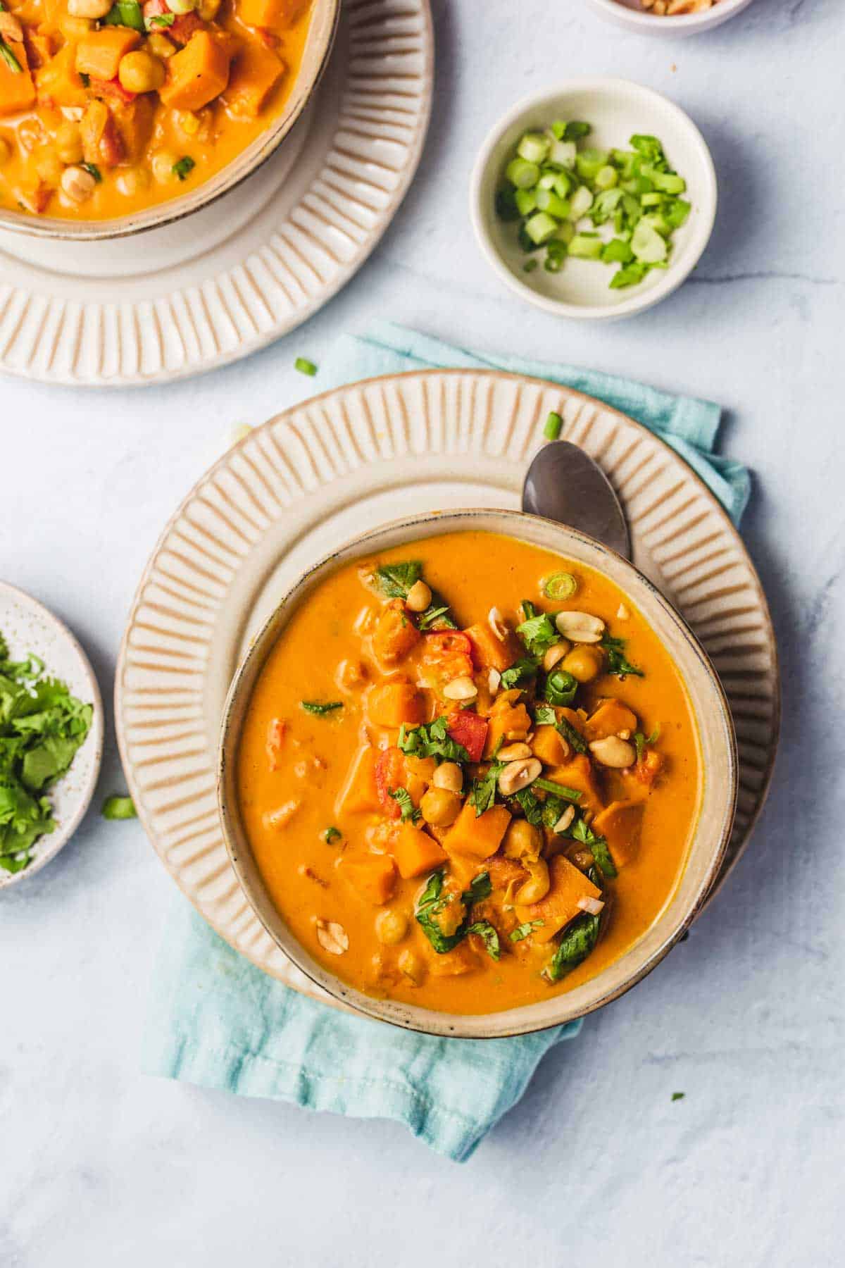 Overhead view of a bowl of sweet potato peanut soup on a plate with a spoon tucked under. Pinch bowl of garnishes off to the side.