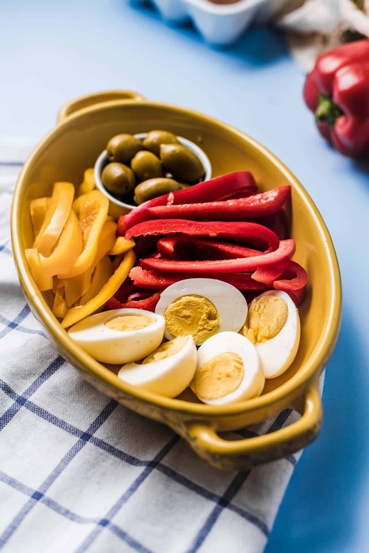 Close up of hard boiled egg snack in a yellow shallow bowl with sliced bell peppers and olives in a white ramekin.
