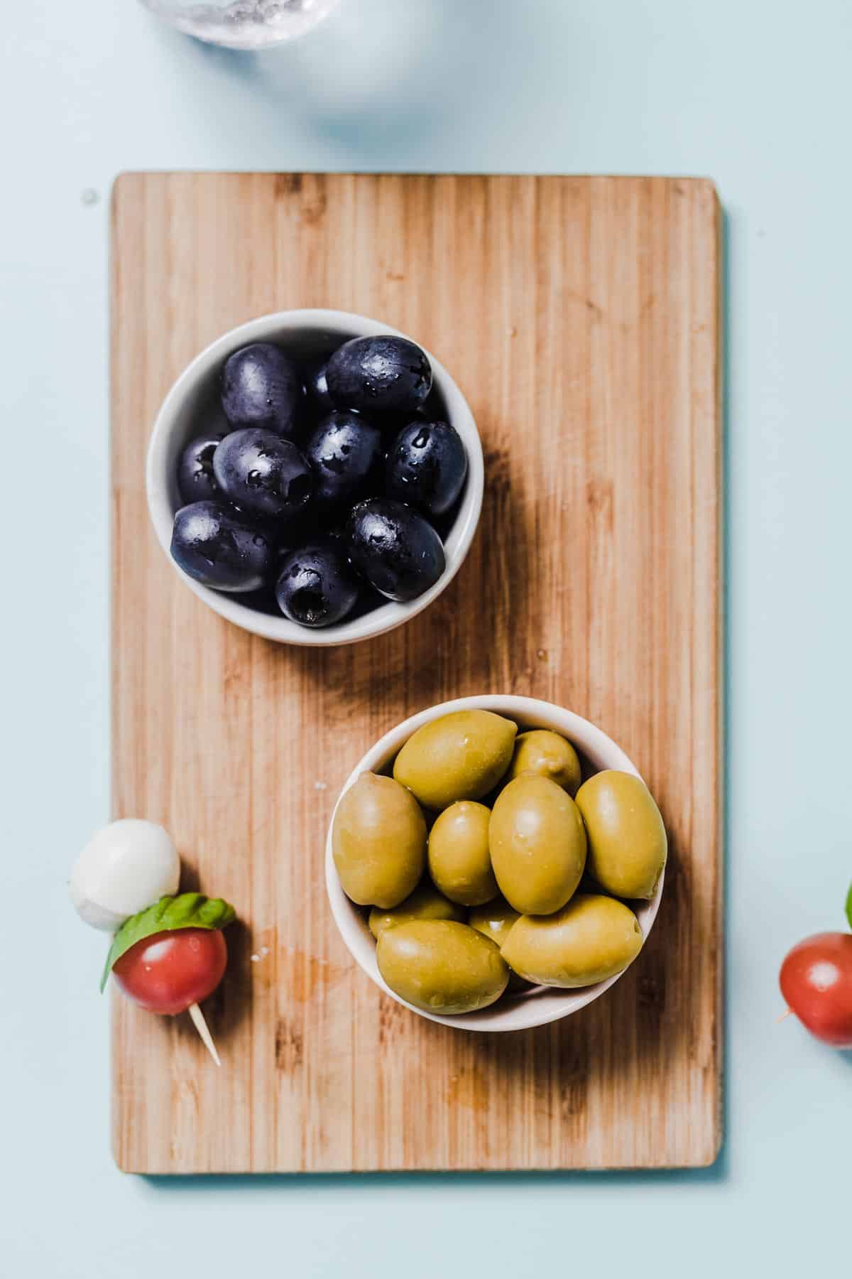 Birds eye view of green and black olives portioned in white bowls on a wooden cutting board.