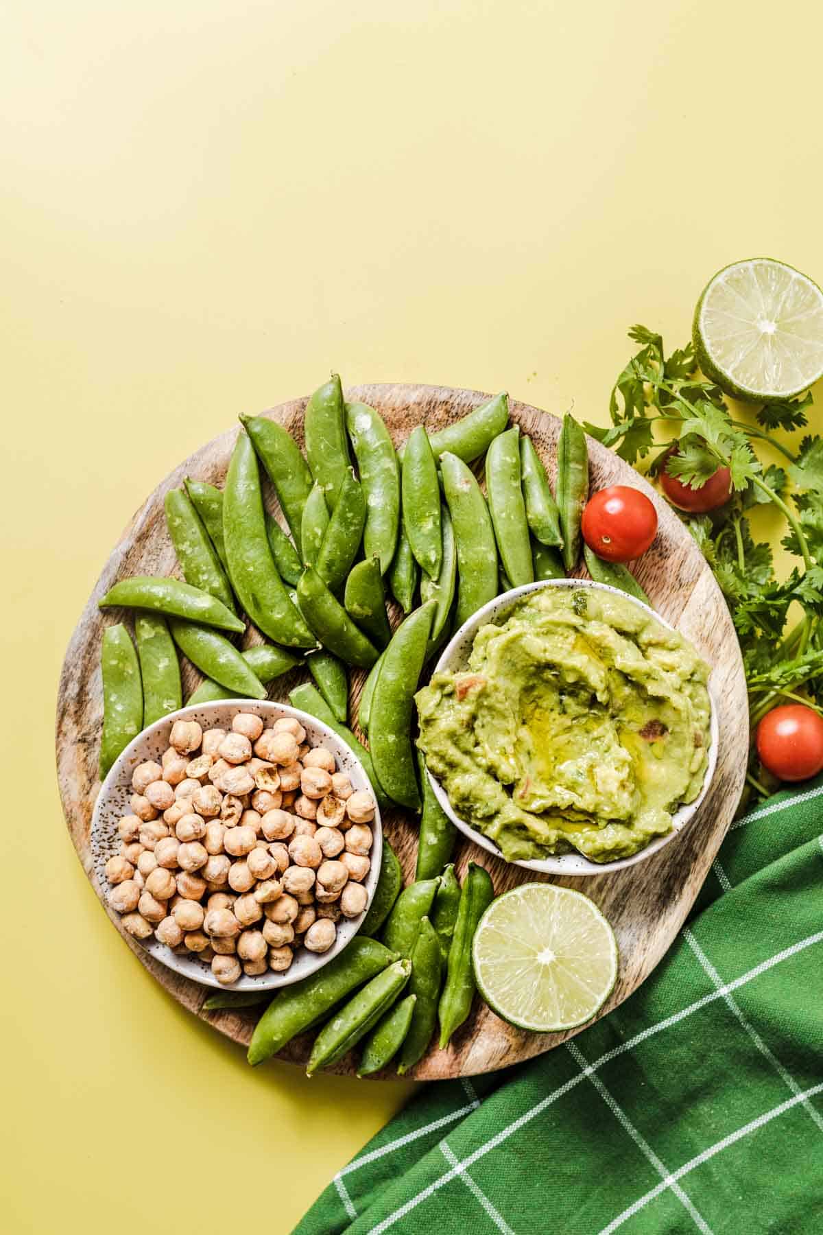 Birds eye view image of sugar snap pea snack on a wooden plate with roasted chickpeas in a white bowl and guacamole in another white bowl.