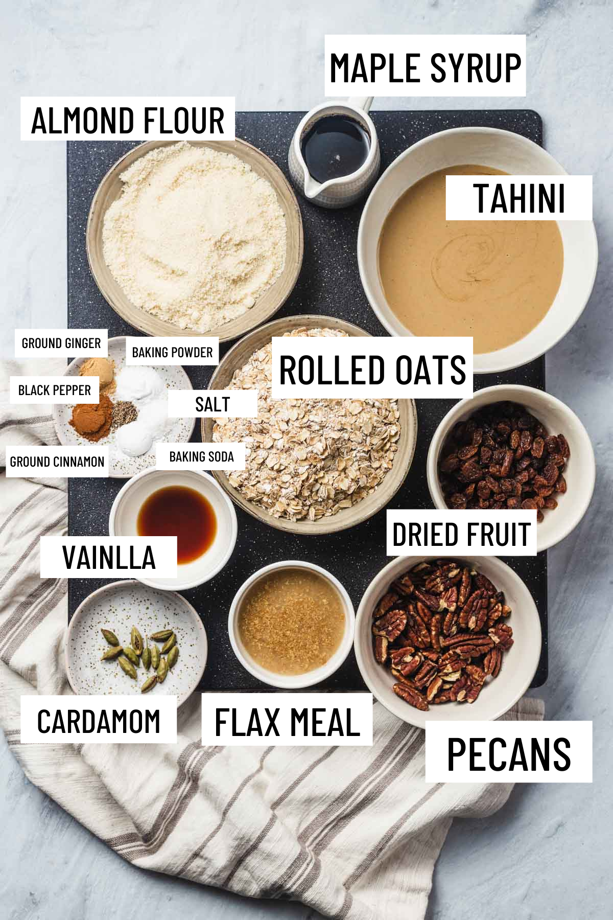 Birds eye view of portioned recipe ingredients including almond flour, maple syrup, tahini, ground ginger, black pepper, ground cinnamon, baking powder, salt, baking soda, vanilla, rolled oats, dried fruit, cardamom, flax meal, and pecans.