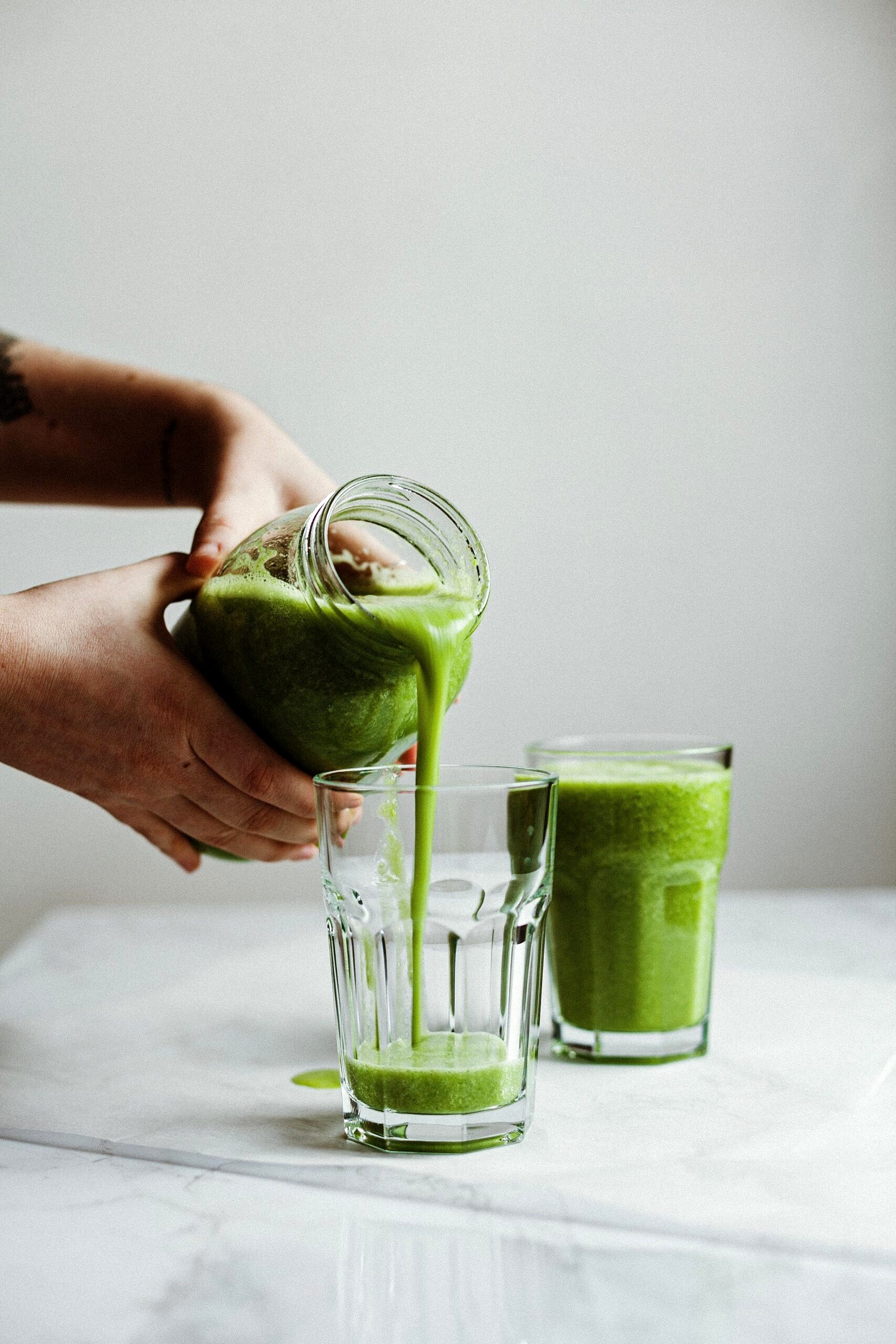 Hands pouring green juice into a clear glass.