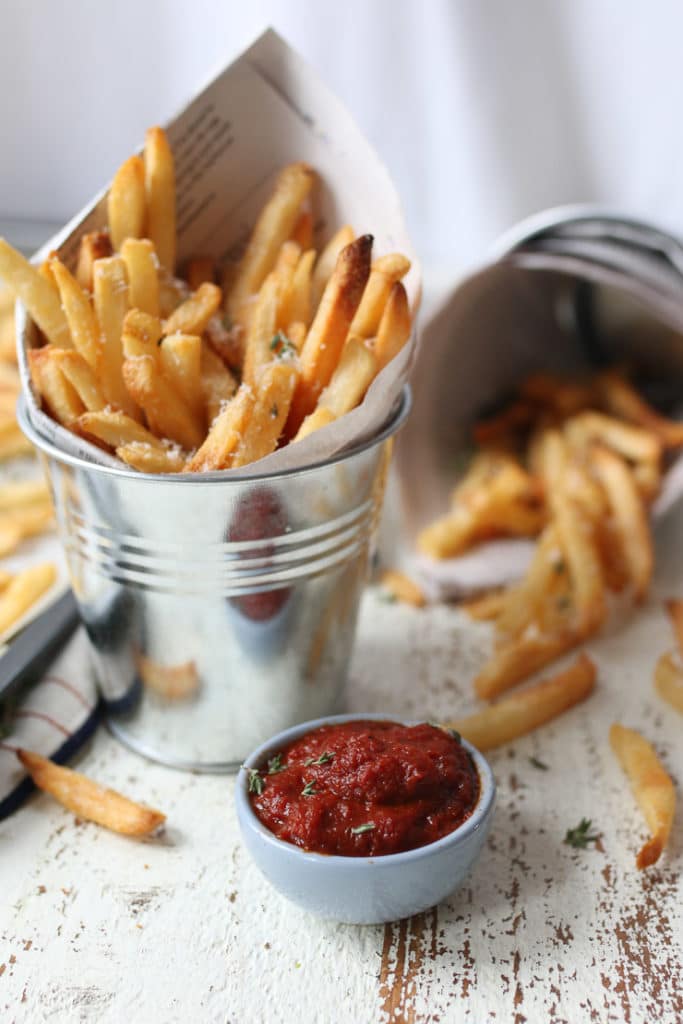 Full shot of a small bowl of ketchup with a tin of french fries in the background. 