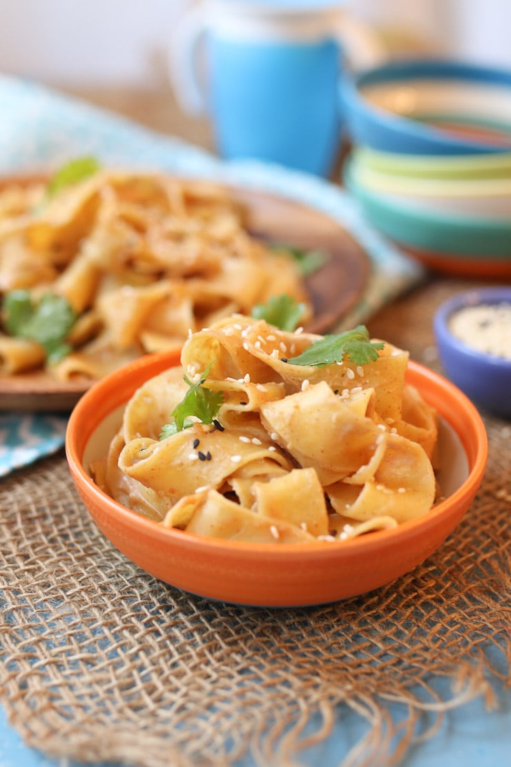 Close up view of the peanut butter sesame noodles in an orange bowl with more noodles on a plate in the background.