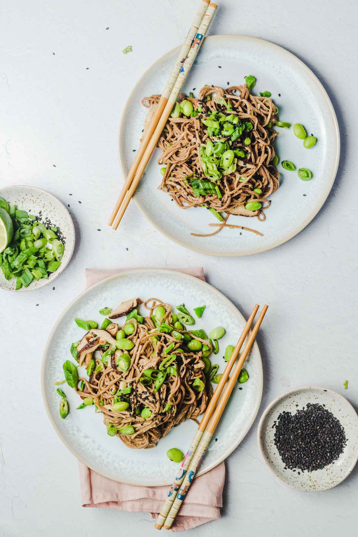 Birds eye view of 2 dishes of miso pasta on white plates with chopsticks resting on each plate. There is a bowl of scallions to the left and plate of black sesame seeds to the right.